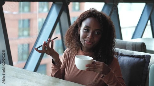 A very beautiful African American woman with long hair listens to a voice message on a smartphone and drinks tea or coffee while sitting at a table in a cafe in the dining room. The woman put her
