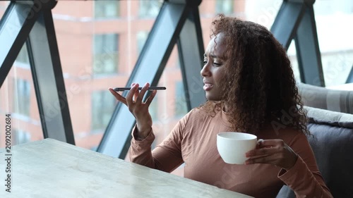 A very beautiful African American woman with long hair listens to a voice message on a smartphone and drinks tea or coffee while sitting at a table in a cafe in the dining room. The woman put her