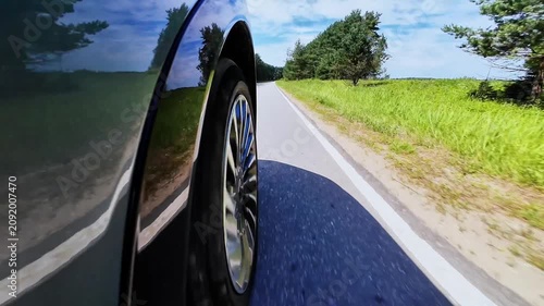 Car tire spinning on asphalt road with green trees alongside and clear blue sky above