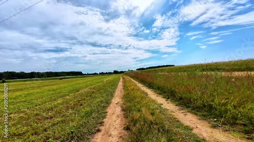 Dirt path dividing lush green fields under a blue sky with scattered clouds