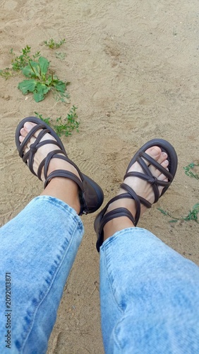 First-person top-down view of feet wearing strappy brown sandals and blue jeans, standing on sandy ground with green weeds.