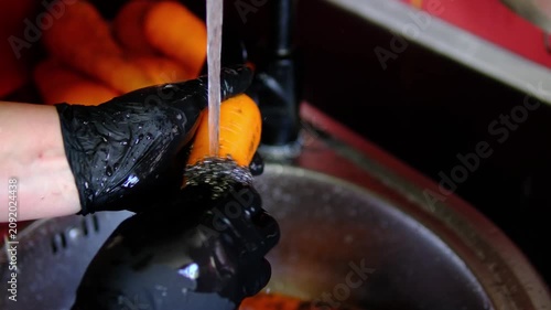 Female hands in black kitchen gloves washing fresh carrots with metal scrubber under running water, cleaning root vegetables from soil before cooking, healthy food preparation at home