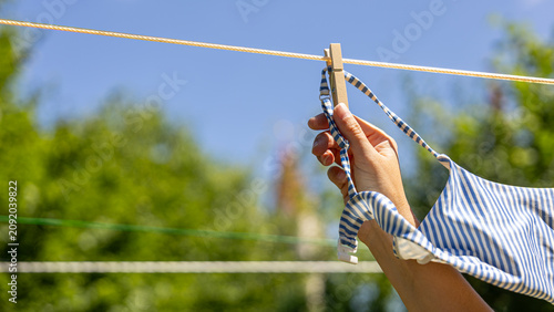 Hand hanging a striped swimsuit on an outdoor clothesline after swimming on a sunny summer day. Eco-friendly air drying, energy saving, sustainable living, summer holidays and local tourism