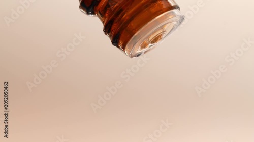 Essential oil slowly dripping from a glass brown bottle, close-up, beige background.