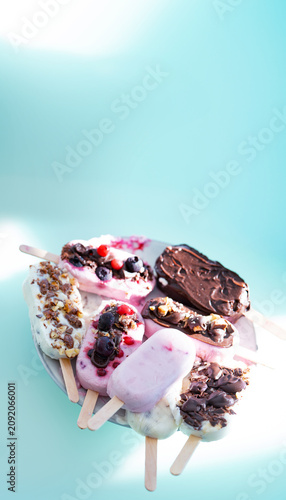 Colorful assortment of homemade yogurt popsicles. Vanilla and chocolate decorated with nuts and frozen berries on ice blue background. Perfect for summer children's birthday party. Vertical overhead c