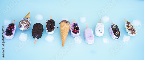 Colorful row of homemade yogurt and milk ice pops. Vanilla and chocolate decorated with nuts and frozen berries with ice cubes on bright blue background. Perfect for a summer children's birthday party