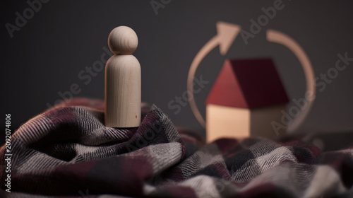 A solitary wooden peg doll sits on a checkered blanket, with a miniature house and an arrowed circular symbol in the background, suggesting themes of home and cycles.