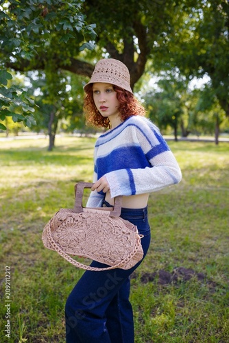 Teenage redhead girl in hat and knit blue striped crop sweater, holds whickered beige purse. Model posing outdoors. Fashion clothing and accessories