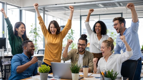 Diverse coworkers cheer with raised fists around a laptop in a bright modern office, suggesting teamwork success for business, startup, or workplace culture campaigns.