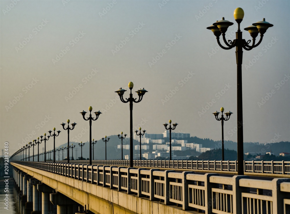 Obraz premium Long concrete bridge with repeating ornate street lamps crosses water. Buildings visible on distant tree covered hill. Daytime scene with clear sky.