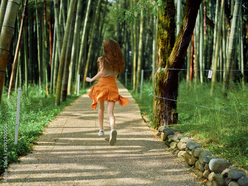 Naklejka premium Woman running on a gravel path through a bamboo forest, back view in an orange skirt, summer sunlight filtering between trunks, motion and freedom on a peaceful park walkway.
