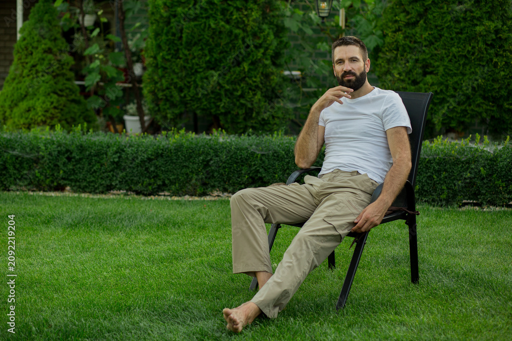 Fototapeta premium Confident bearded man sitting on outdoor chair in a green backyard garden, looking at camera with a thoughtful expression. Relaxed lifestyle, leisure time, personal freedom, mindfulness