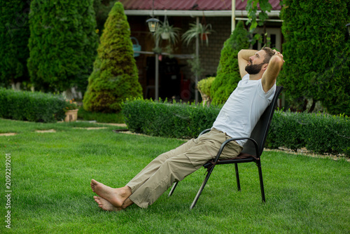 Bearded man relaxing on outdoor chair in a green backyard garden after working remotely. Freelance professional taking a break, enjoying fresh air, work-life balance, stress-free lifestyle