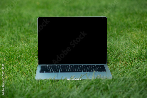 Modern laptop with blank black screen placed on green grass outdoors. Mockup template for website, software, online business, remote work, digital marketing, presentation or advertising design.