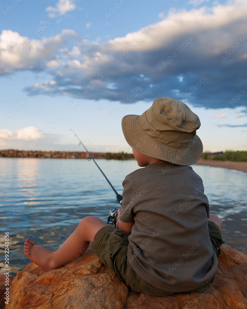 Fototapeta premium Young Boy Joyfully Casting His Line from Tranquil Harbor at Sunset, Embracing the Peace of Nature