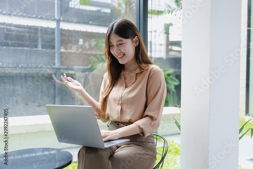 Smiling young woman using a laptop by a large window in a bright cafe or office, working online in a relaxed modern space with natural light and a cheerful mood.