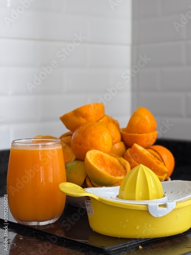 A commercial eye-level photograph of a yellow and white plastic manual citrus juicer placed in the center of many fresh orange halves, creating a neat pattern on a black reflective kitchen counter. Th