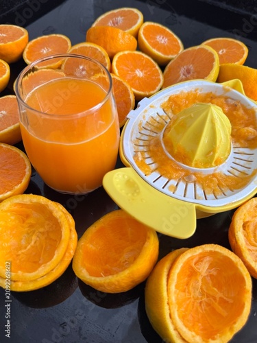 A commercial eye-level photograph of a yellow and white plastic manual citrus juicer placed in the center of many fresh orange halves, creating a neat pattern on a black reflective kitchen counter. Th