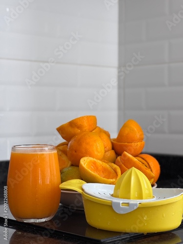 A commercial eye-level photograph of a yellow and white plastic manual citrus juicer placed in the center of many fresh orange halves, creating a neat pattern on a black reflective kitchen counter. Th