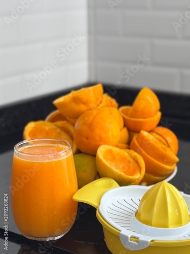 A commercial eye-level photograph of a yellow and white plastic manual citrus juicer placed in the center of many fresh orange halves, creating a neat pattern on a black reflective kitchen counter. Th