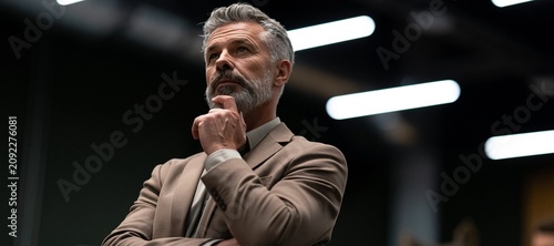 Mature businessman with gray hair and beard thoughtfully looking up and touching his chin in a dimly lit modern office environment