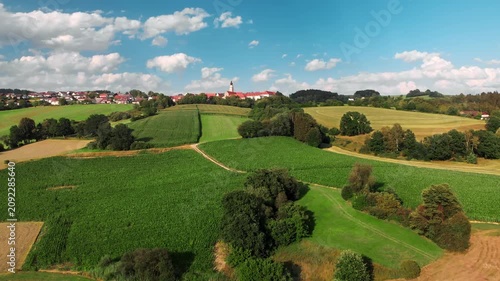 Flying towards a village on the horizon on beautiful green hills in Bavaria, Germany, with trees, blue sky and fluffy white clouds in the background