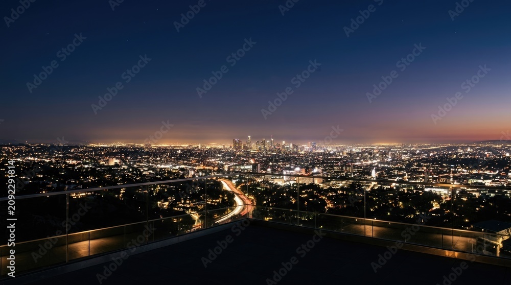 Fototapeta premium A person stands on a balcony overlooking a sprawling cityscape at night. The dark sky is illuminated by the lights of Los Angeles, stretching out towards the horizon.