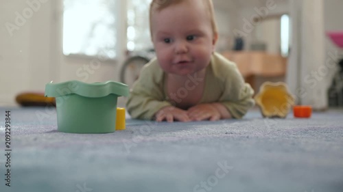 Low-angle, shallow-focus view of an infant crawling across a carpeted living room floor toward scattered plastic stacking cups and toys