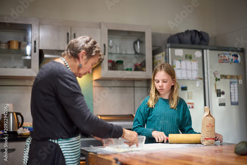Grandmother and granddaughter baking together in a cozy kitchen while the girl rolls dough with a rolling pin