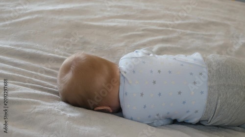 The baby rolls over from his tummy to his back. The baby is practicing rolling over on the bed, looking at the camera. The baby is radiating positive emotions