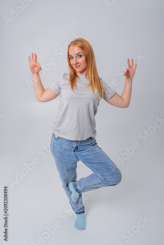 Woman standing on one leg, balancing while smiling at the camera and holding up three fingers on one hand and making an OK gesture with the other hand