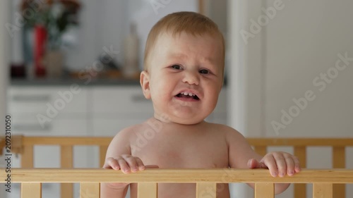 The baby peers over the edge of the crib, displaying a variety of facial expressions, from curiosity to pity, in the cozy and welcoming atmosphere of the home