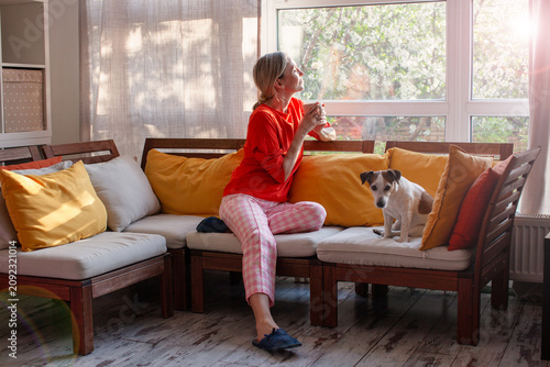 Quadro su tela Woman sitting on a sofa in a living room holding a cup of coffee and looking out a window