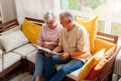 Elderly woman and man looking at family photo album on couch. Senior couple sitting together at home remembering past memories. Nostalgia and love in retirement lifestyle concept.