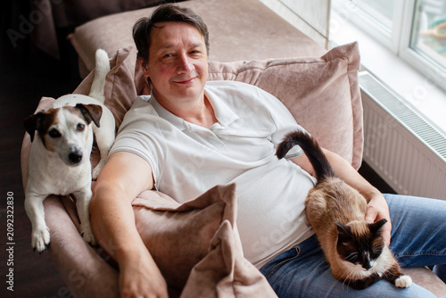 Man relaxing on a sofa with his pet cat and dog at home. Mature guy spending leisure time together with domestic animals. Friendship between human and pets, animal care concept.