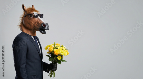 A man in a horse mask and business suit wearing sunglasses holding a yellow flower bouquet, studio