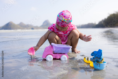 Adorable Asian Toddler Girl Playing with Beach Toys on the Shore,A cute little Asian toddler girl wearing a pink swimsuit and swimming cap, playing with a pink toy truck and sand bucket on a beautiful