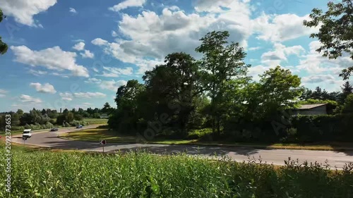 Time lapse of cars driving on a curved country asphalt road next to green hills and trees under a dynamic blue sky with moving white clouds