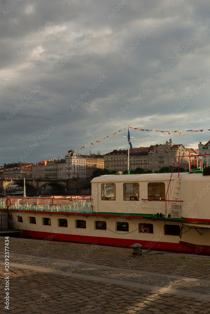 Obraz premium A moored riverboat with a white and red hull sits alongside a stone quay in front of a row of historic multi-story buildings. Festive bunting stretches above