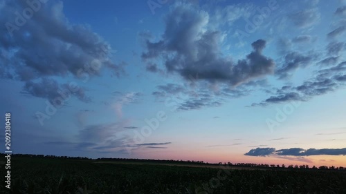 Time lapse of beautiful sunset sky with colorful gradients and dark cumulus clouds moving smoothly above a vast green agriculture field at evening twilight