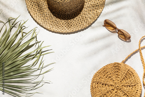 Straw hat, sunglasses, woven bag, and palm leaf rest on a sunny white surface. Summer vacation aesthetic, tropical travel vibe, sunny beach lifestyle, summer holiday. Flat lay banner with copy space