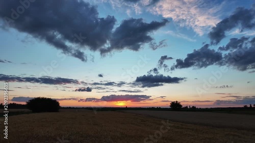Time lapse of a vibrant sunset with orange glow sinking below the horizon of a vast wheat field under a blue sky with moving dark clouds
