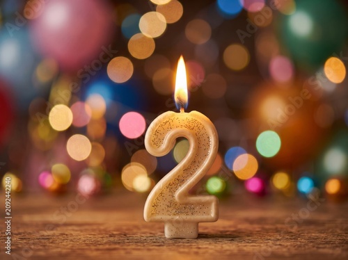 Burning number 2 candle for a second birthday or anniversary celebration with festive colorful bokeh lights and balloons in the background