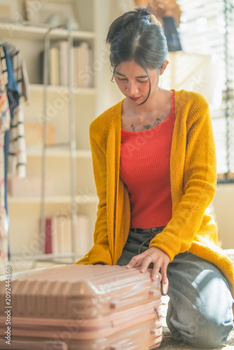Young Asian woman in yellow cardigan packing suitcase with folded clothes, sitting on floor in sunlit bedroom, preparing for travel vacation trip, cozy home lifestyle morning routine scene, warm light