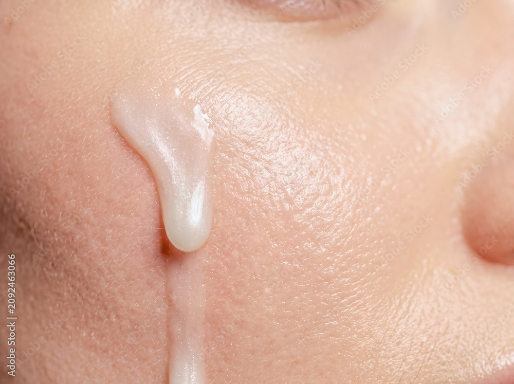 Fototapeta premium Macro close up of a white cosmetic cream lotion drop dripping down a smooth clean woman cheek skin surface. Skincare product application and moisturizing beauty concept.