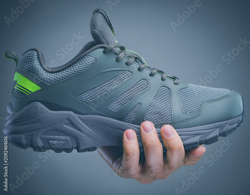 Close-up of a gray athletic shoe with neon green accent on heel, held in hand, against blue gradient, showcasing product in studio setting