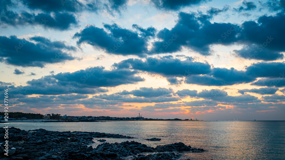 Obraz premium Dramatic dark clouds over a calm ocean and rocky volcanic coastline during a beautiful colorful sunset.