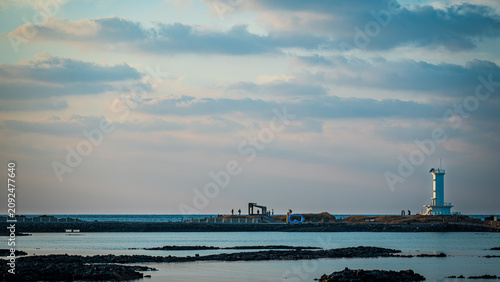 Schilderij op canvas Scenic view of a white lighthouse and distant silhouetted people on a rocky coastal breakwater under a soft cloudy sky at dusk