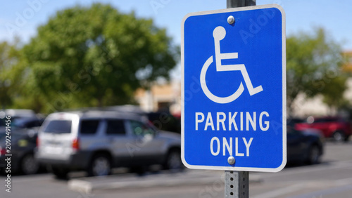 Close-up view of blue and white handicap parking sign in shopping center parking lot on sunny day with blurred cars, accessibility concept, public facility photography, blurred