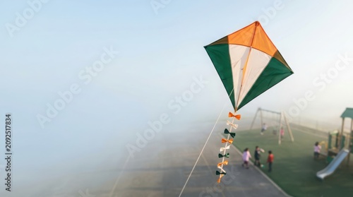Colorful indian flag inspired kite with ribbon tail flying high over playground area filled with playing children in mist.